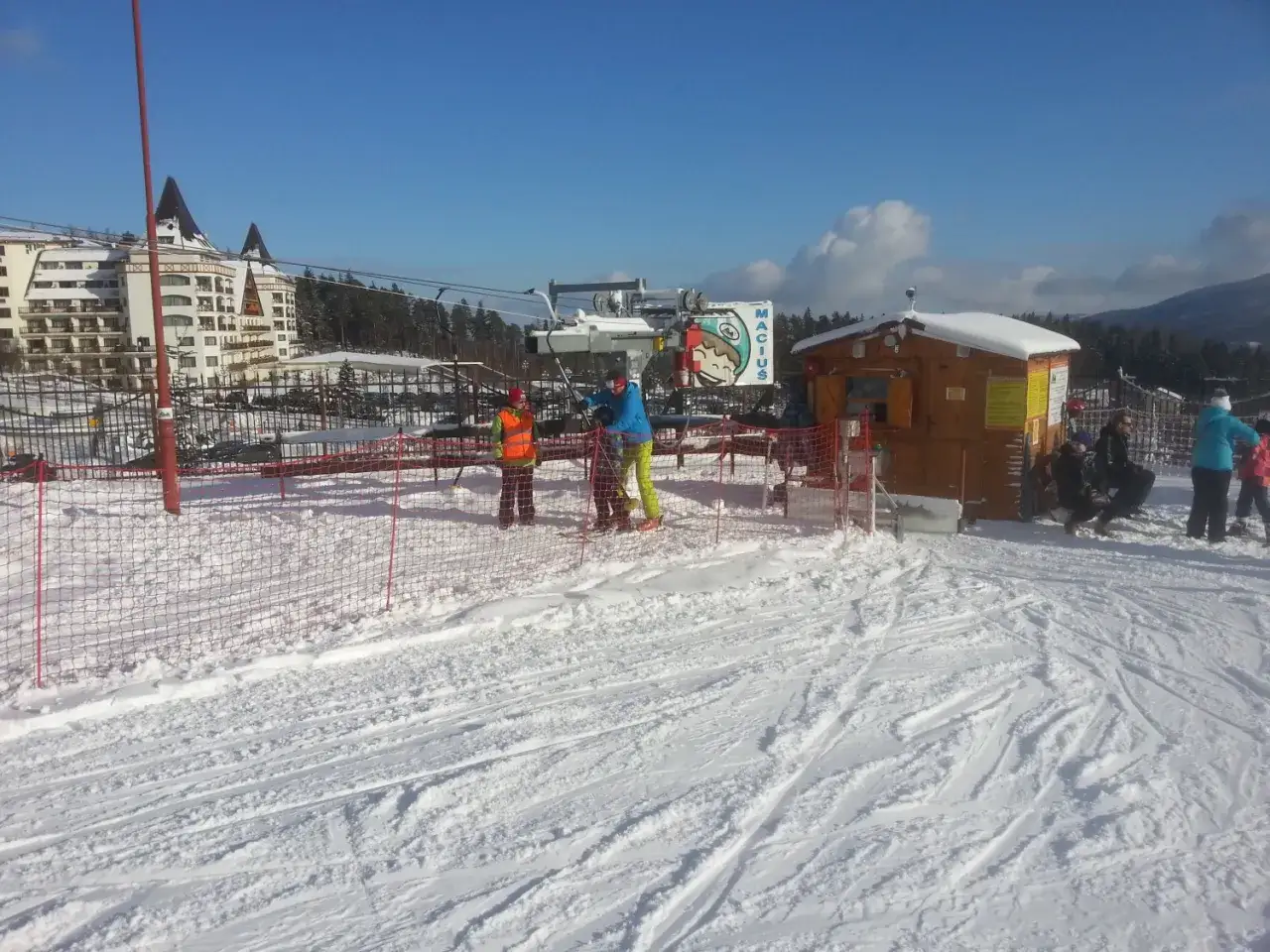 Stok Macius Karpacz zimą. Narciarze czekają na wyciąg przy drewnianej budce. W tle hotel i góry.