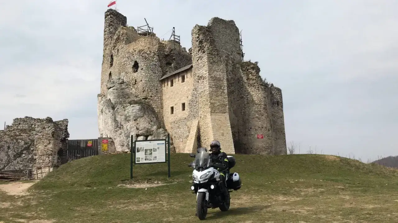 Motocyklista na tle ruin zamku na Szlaku Orlich Gniazd. Biały motocykl z kuframi, w tle kamienne mury i polska flaga.