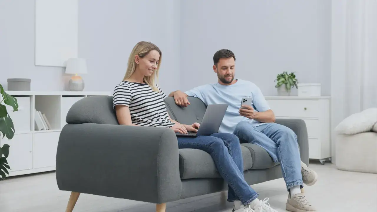Para planuje zamianę domu na mieszkanie, przeglądając oferty na laptopie i telefonie, siedząc na kanapie.