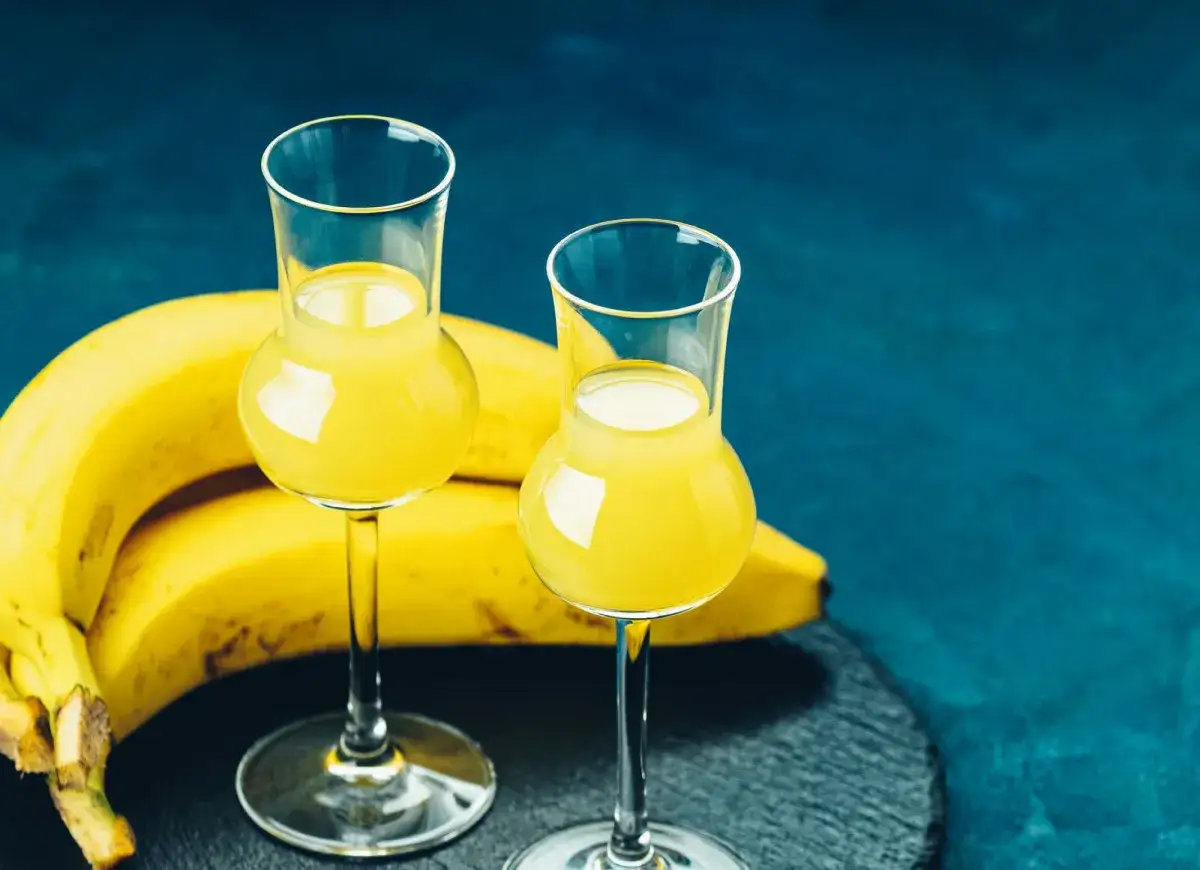 Dwa kieliszki z żółtym likierem bananowym, obok pęk bananów. Idealny przepis na domowy likier bananowy.