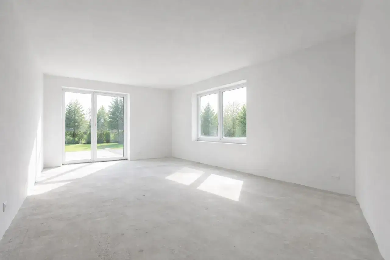 Puste pomieszczenie z betonową podłogą i białymi ścianami. Duże okna wychodzą na zielony ogród. Idealne miejsce, by zastanowić się, czy kłaść gładź pod tapetę.