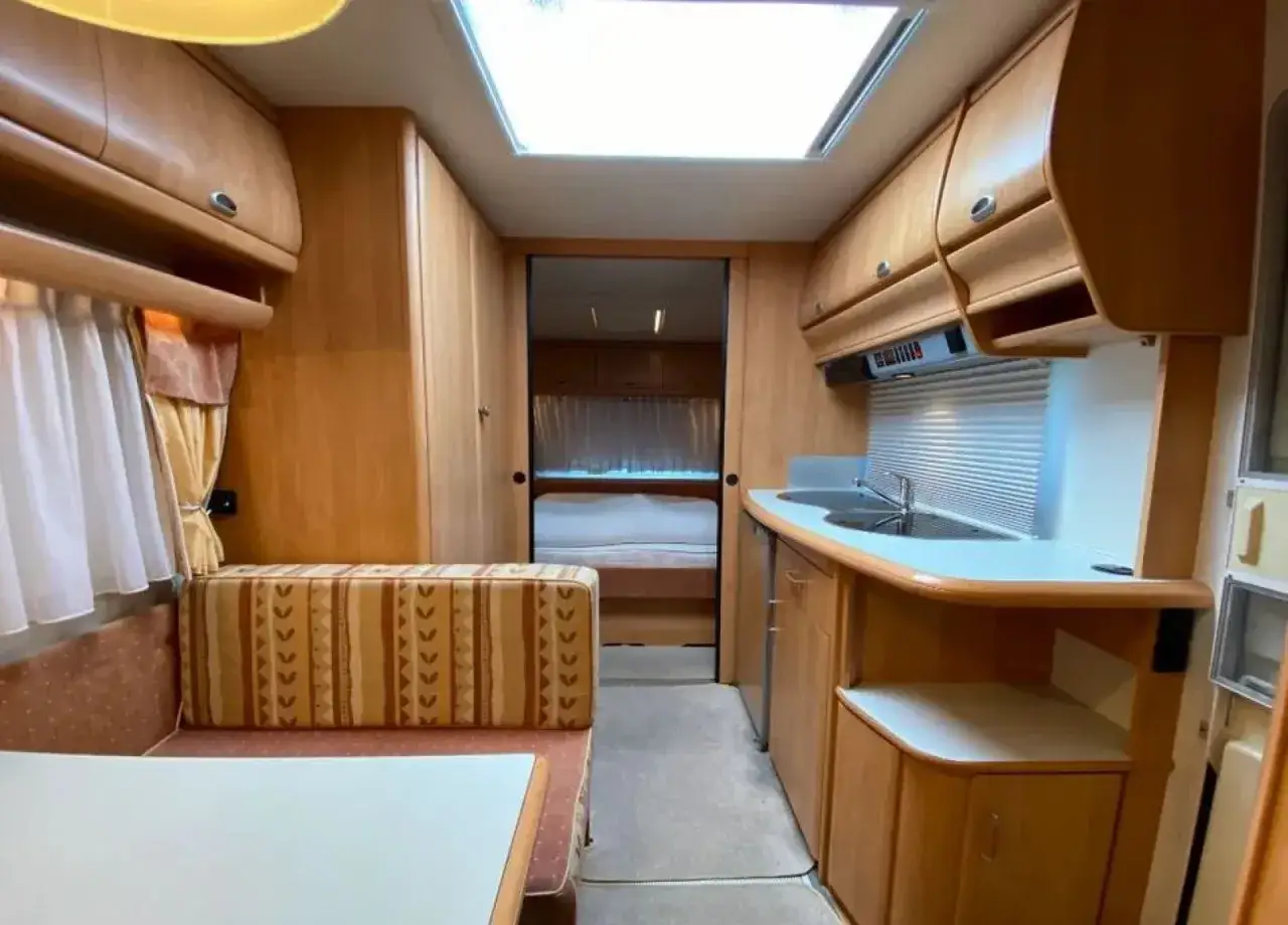 Wnętrze kampera Hobby Prestige 495: kuchnia z blatem, zlewem i szafkami, kanapa z wzorzystymi poduszkami, st&oacute;ł i widok na ł&oacute;żko.