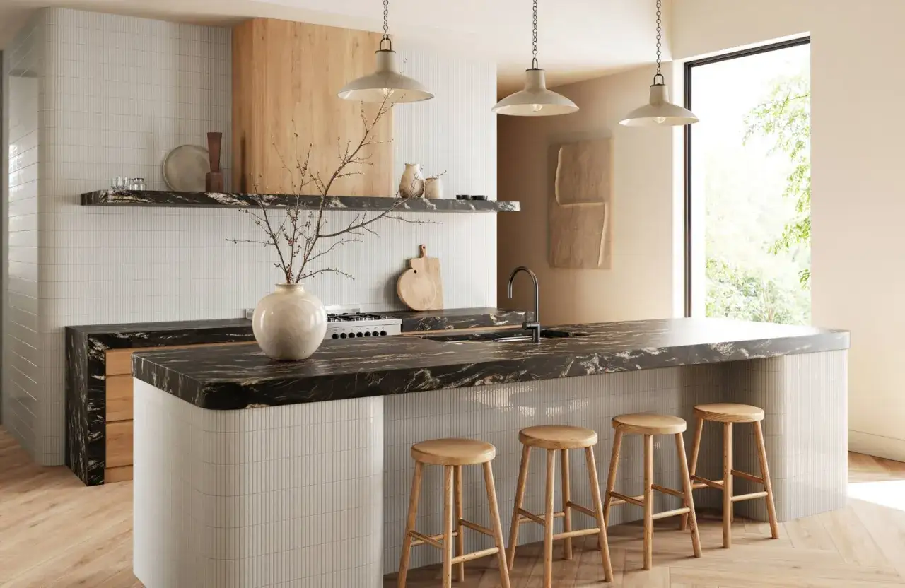 Nowoczesna kuchnia z wyspą, czarnym blatem i drewnianymi stołkami. Oświetlenie og&oacute;lne tworzy przytulną atmosferę.