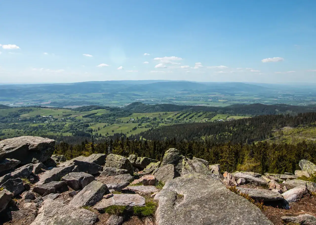 Widok z kamienistego szczytu na zielone doliny i wzg&oacute;rza. W oddali widać pasma g&oacute;rskie. To krajobraz z wieży widokowej Tr&oacute;jmorski Wierch.