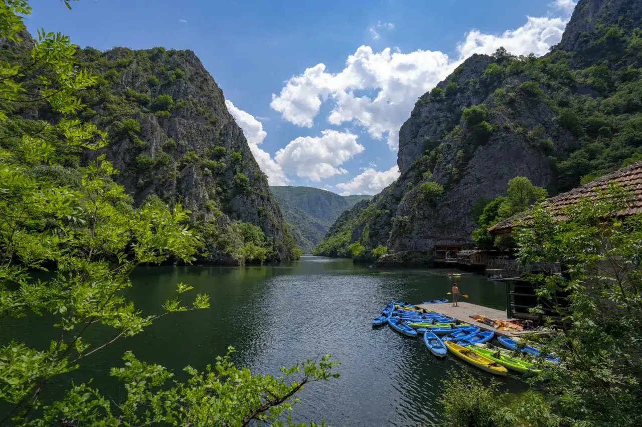 Wodospady i skaliste zbocza kanionu Matka, z rzędem kolorowych kajak&oacute;w przy drewnianym pomoście.