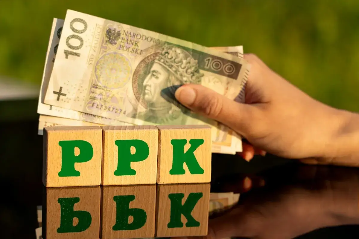 Dłonie trzymają polskie banknoty nad klockami z napisem PPK. Zmiana pracy może oznaczać nowe możliwości oszczędzania.