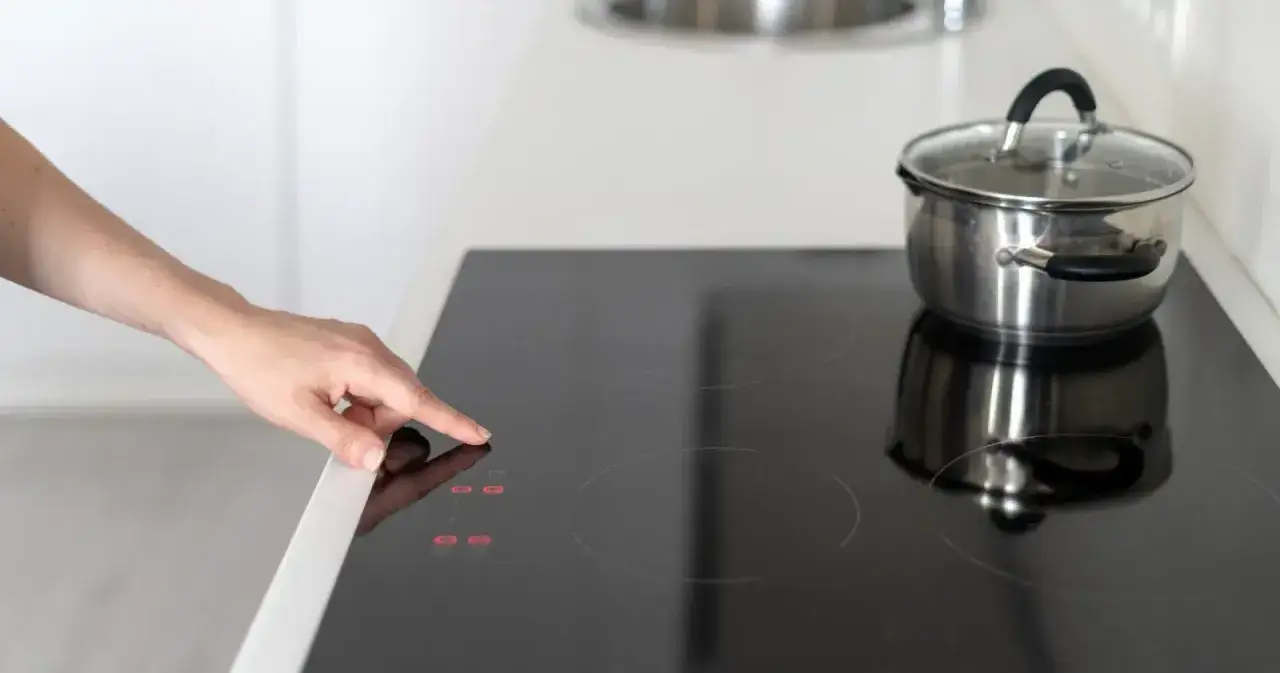 Dłoń naciska przycisk na płycie indukcyjnej, gdzie garnek czeka na gotowanie. 