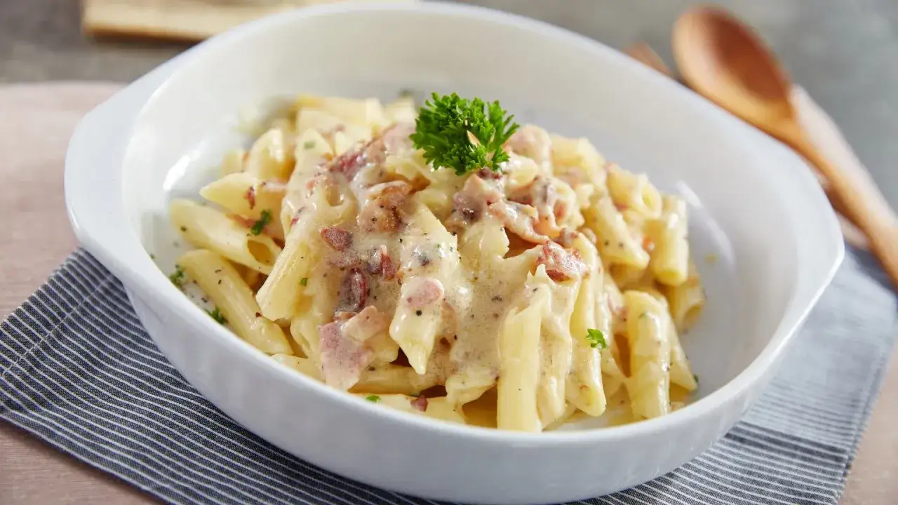 Penne rigate w kremowym sosie z chrupiącym boczkiem i natką pietruszki, podane w białej misce. Pyszny makaron z boczkiem.
