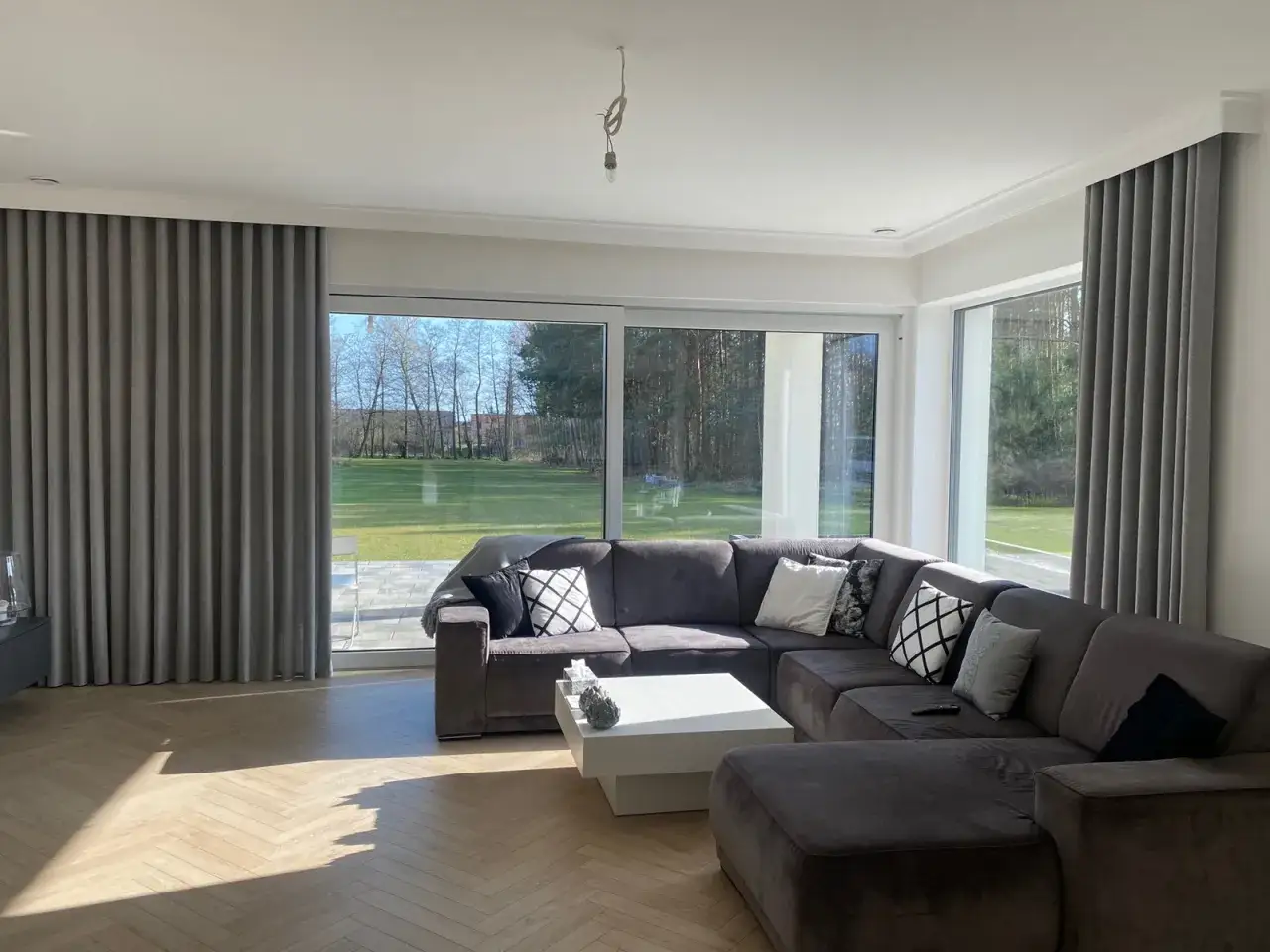 Nowoczesny salon z szarą kanapą i zasłonami, inspiracje szare ściany. Duże okna wychodzą na zielony ogr&oacute;d.