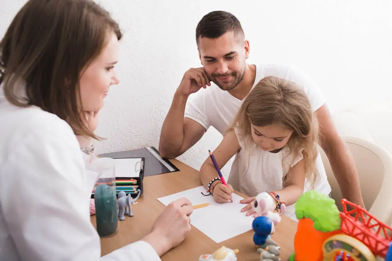 Ojciec i córka na spotkaniu z terapeutką. Dziecko rysuje, a opinia z poradni pomoże w dalszej terapii.