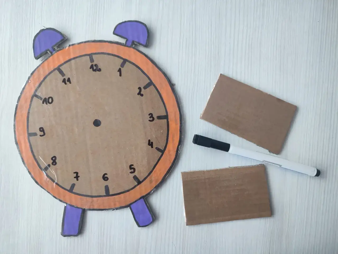 Zrób własny zegar z tektury! Na stole leży gotowa tarcza zegara, dwa kawałki tektury i pisak.