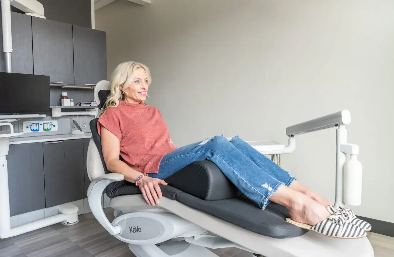 Uśmiechnięta kobieta w jeansach i bluzce siedzi na fotelu dentystycznym, gotowa na wizytę u dentysty.