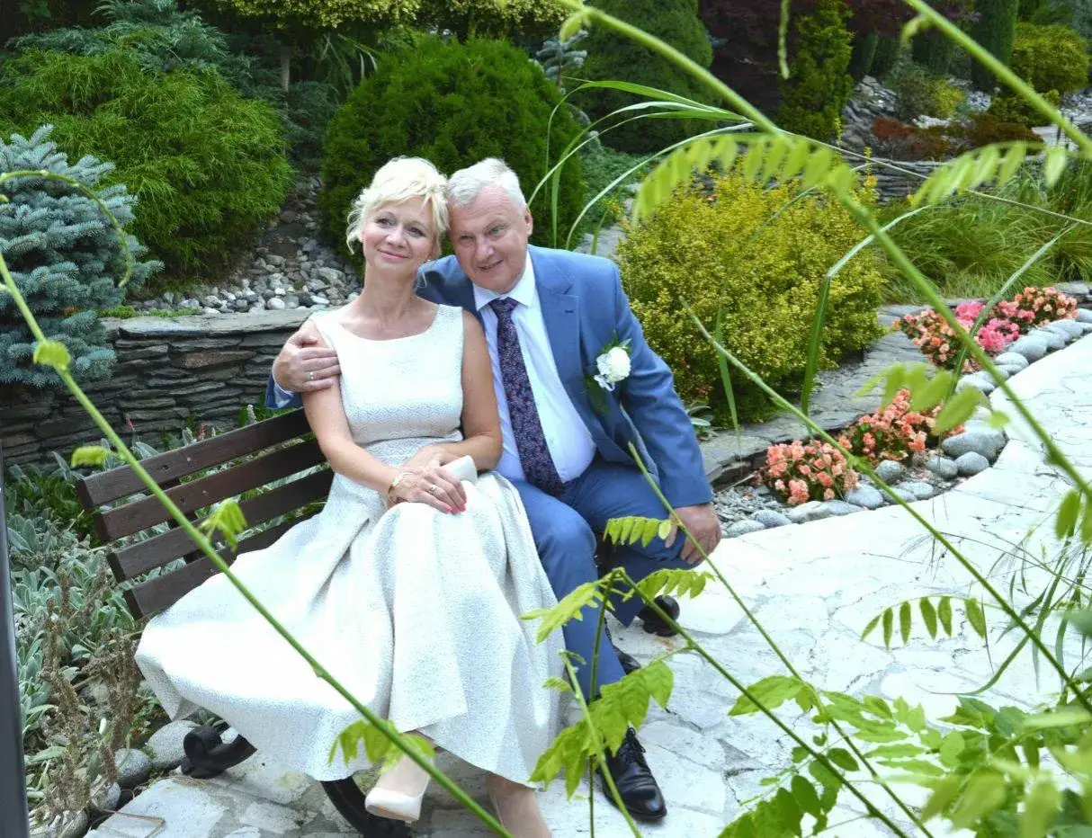 Para 50-latki sukienki do ślubu cywilnego. Para siedzi na ławce w ogrodzie, ona w eleganckiej, jasnej sukience, on w niebieskim garniturze.