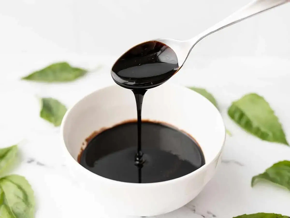 Gęsty, ciemny sos balsamiczny spływa z łyżki do białej miseczki. Wokół zielone listki bazylii.