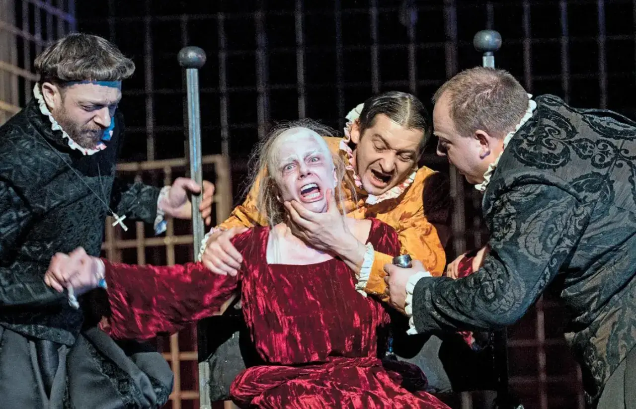 Aktorzy w strojach z epoki odgrywają scenę tortur. Kobieta w czerwonej sukni krzyczy, trzymana przez dwóch mężczyzn.