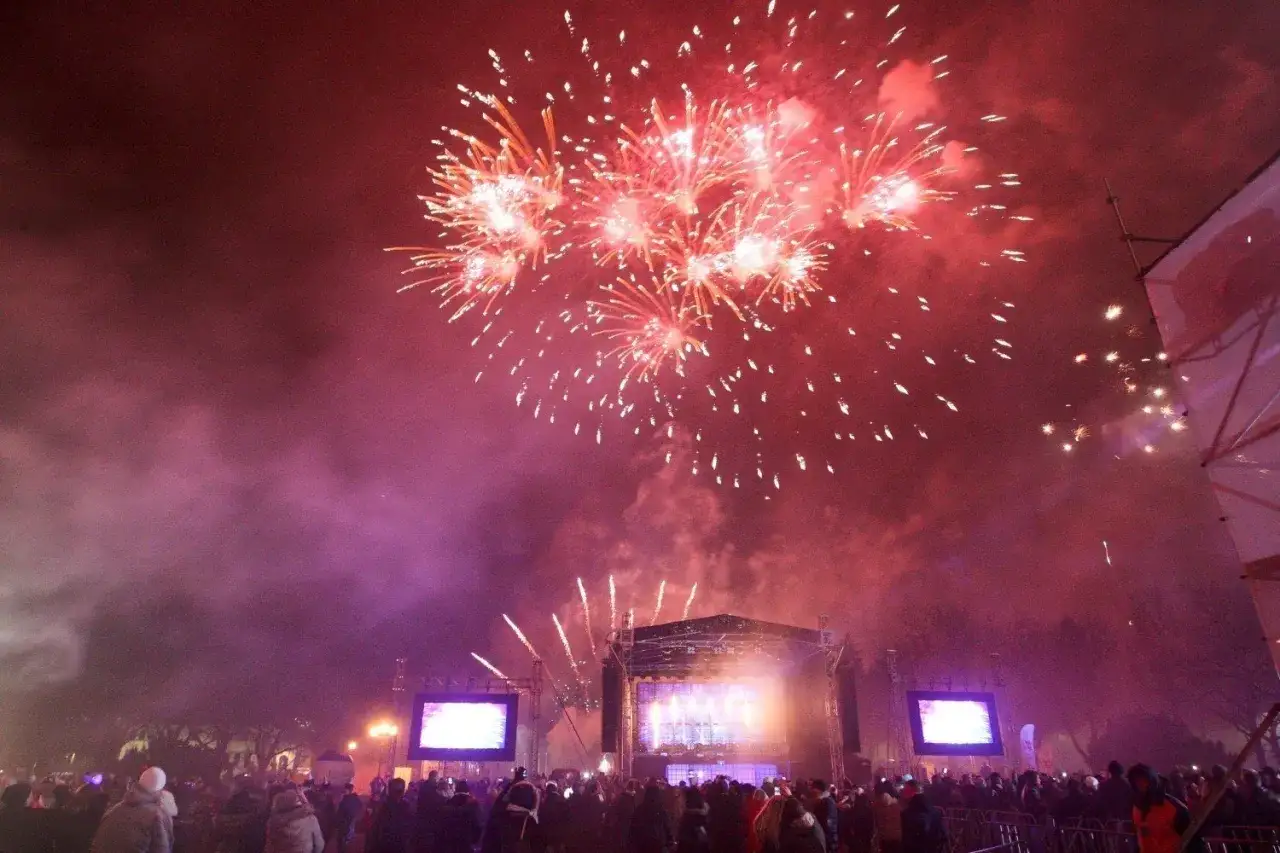 Spektakularny pokaz fajerwerków nad sceną, gdzie odbywa się koncert sylwestrowy w Warszawie. Tłum ludzi świętuje nadejście Nowego Roku.
