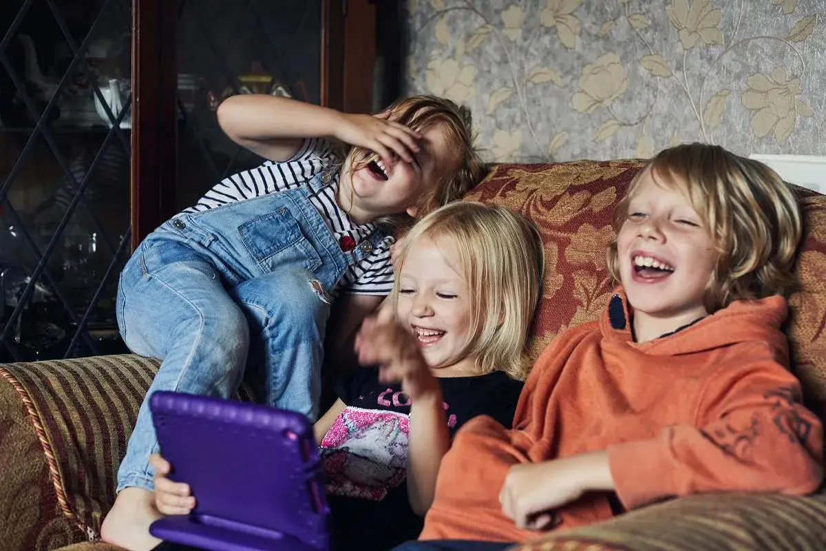 Tr&oacute;jka dzieci śmieje się radośnie, jedno dziecko śmieje się samo do siebie, patrząc na tablet.