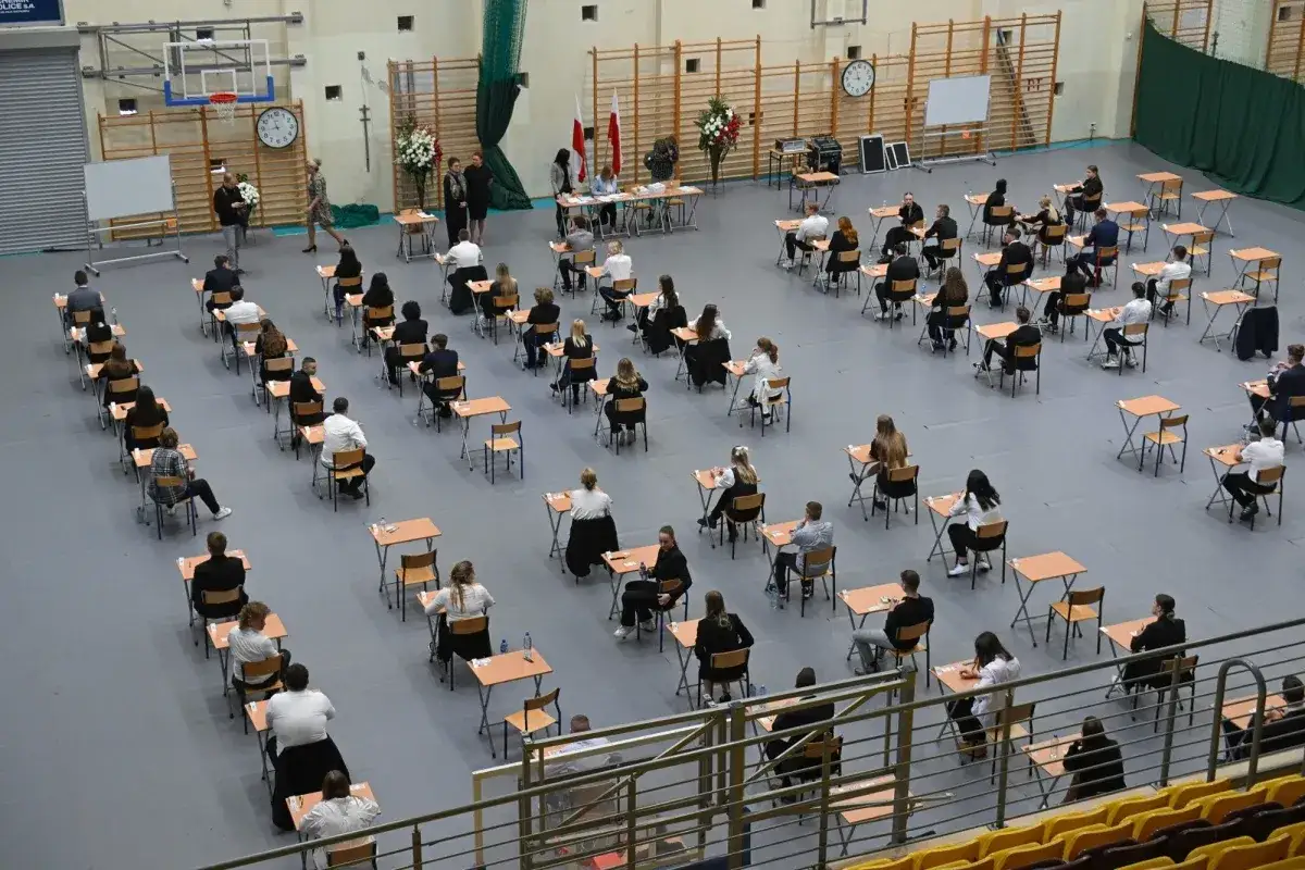 Uczniowie siedzą przy ławkach w sali gimnastycznej, przygotowując się do egzaminu. Ile lat w liceum minęło, by dojść do tego momentu?