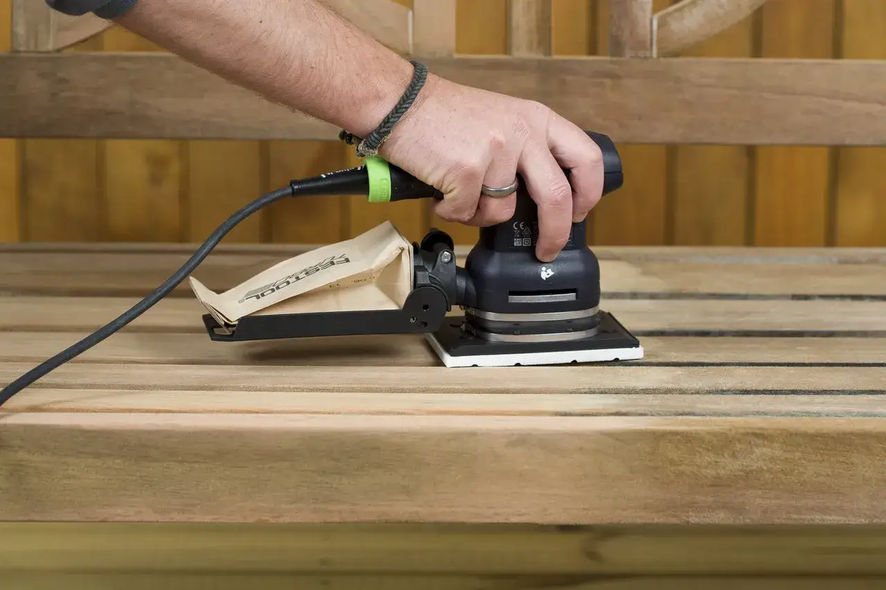 Dłoń trzyma szlifierkę, która pomaga jak usunąć lakier z drewna. Worek na pył zbiera resztki.