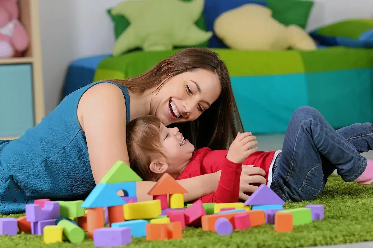 Uśmiechnięta opiekunka bawi się z dzieckiem kolorowymi klockami. Ile bierze opiekunka do dzieci? Tyle radości!