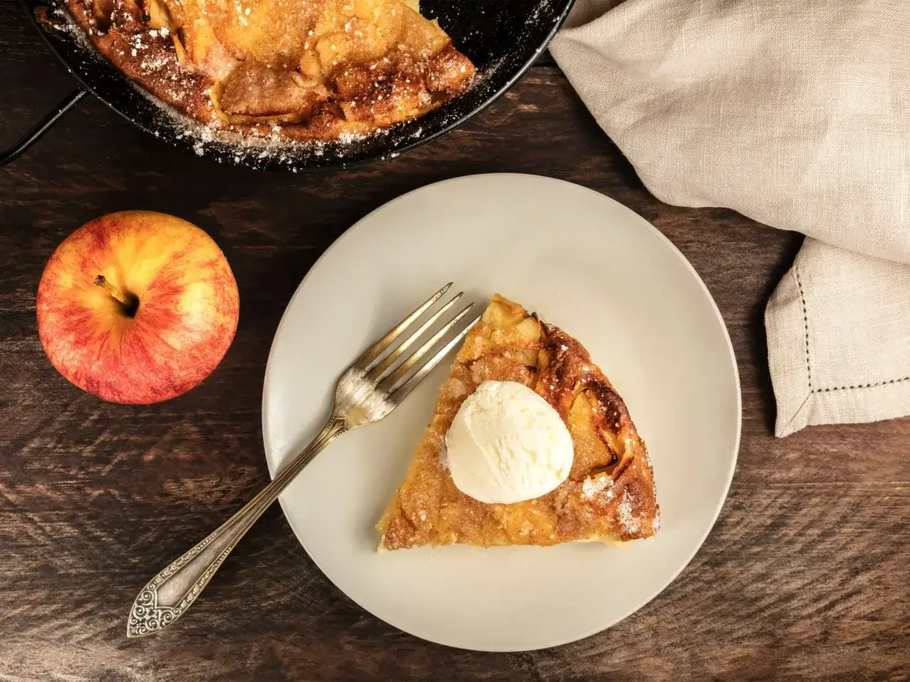 Jak zrobić szarlotka prosty przepis: pyszny kawałek szarlotki z lodami na talerzu, obok jabłko i widelczyk.