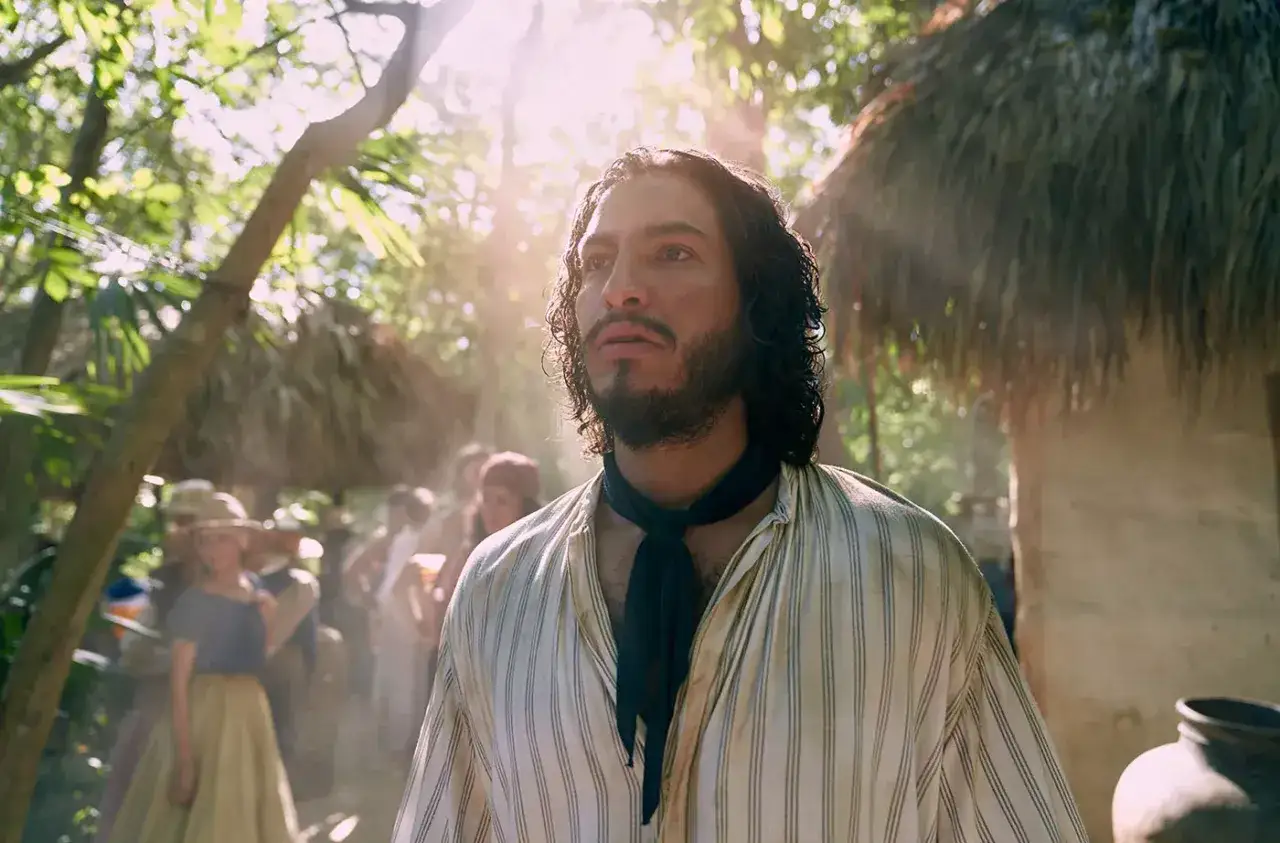 Młody mężczyzna z burzą ciemnych włosów i zarostem, ubrany w pasiastą koszulę, stoi na tle słonecznego lasu i chat krytych strzechą. Scena z serialu 