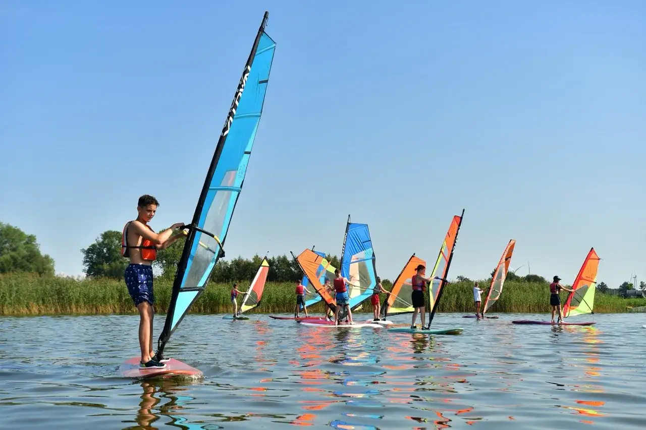 Młodzi ludzie uczą się windsurfingu na spokojnym jeziorze. Czy warto jechać do Dąbk&oacute;w? Zdecydowanie tak, jeśli lubisz wodne sporty!
