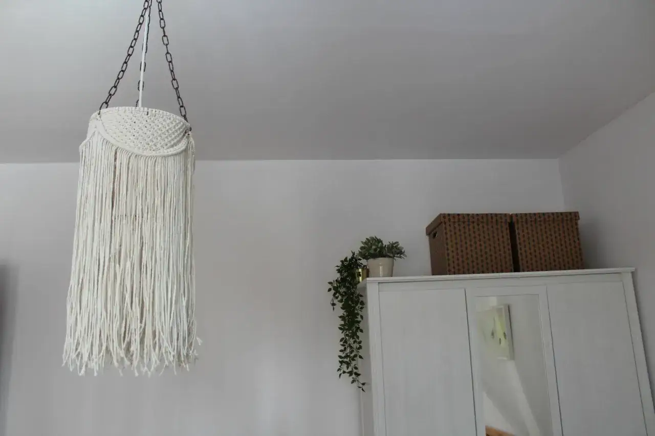 Jak zrobić lampę ze sznurka? Oto inspiracja: makramowa osłona na lampę wiszącą, wisząca na łańcuchu, z frędzlami. Obok szafa z doniczką.