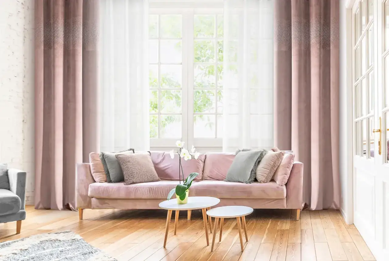 Eleganckie, r&oacute;żowe zasłony z brokatowym wzorem i białe firany tworzą nowoczesne zasłanianie okien. Miękka kanapa i stoliki dopełniają wnętrze.