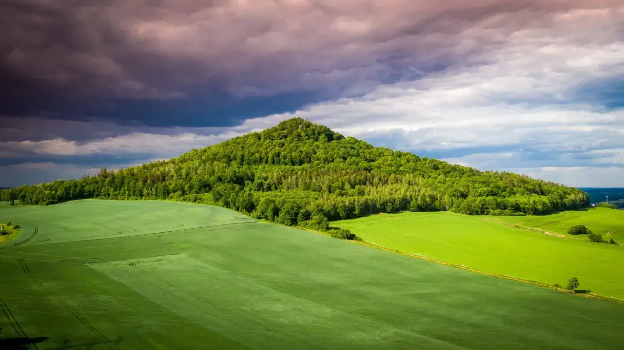 Zielone pola i zalesione wzgórze, przypominające krajobraz krainy wygasłych wulkanów, pod burzowym, fioletowym niebem.