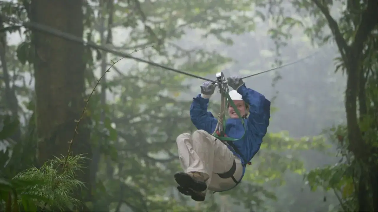 Kobieta w kasku i niebieskiej kurtce zjeżdża na tyrolce przez mglisty las. To prawdziwy **outdoor**!