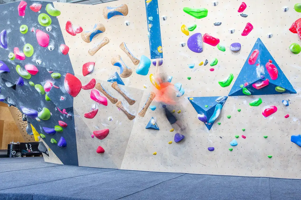 Kolorowa ściana do wspinaczki, gdzie wspinacz w pomarańczowej koszulce pokonuje trudną trasę. To prawdziwa **skala boulderowa** pełna chwyt&oacute;w.