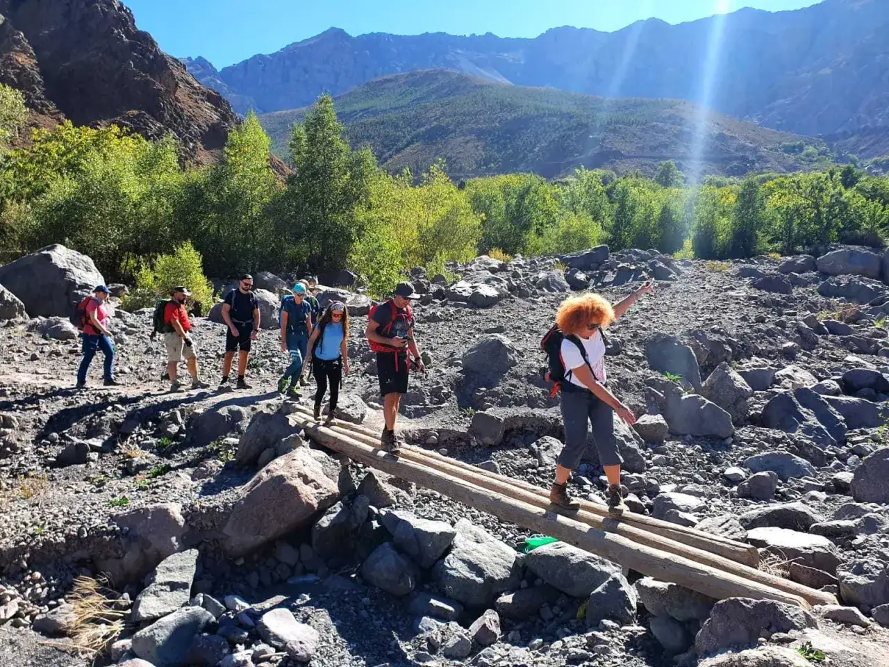 Grupa turyst&oacute;w przemierza kamienisty teren w g&oacute;rach Atlas w Maroku, pokonując strumień po prowizorycznym moście z bali.