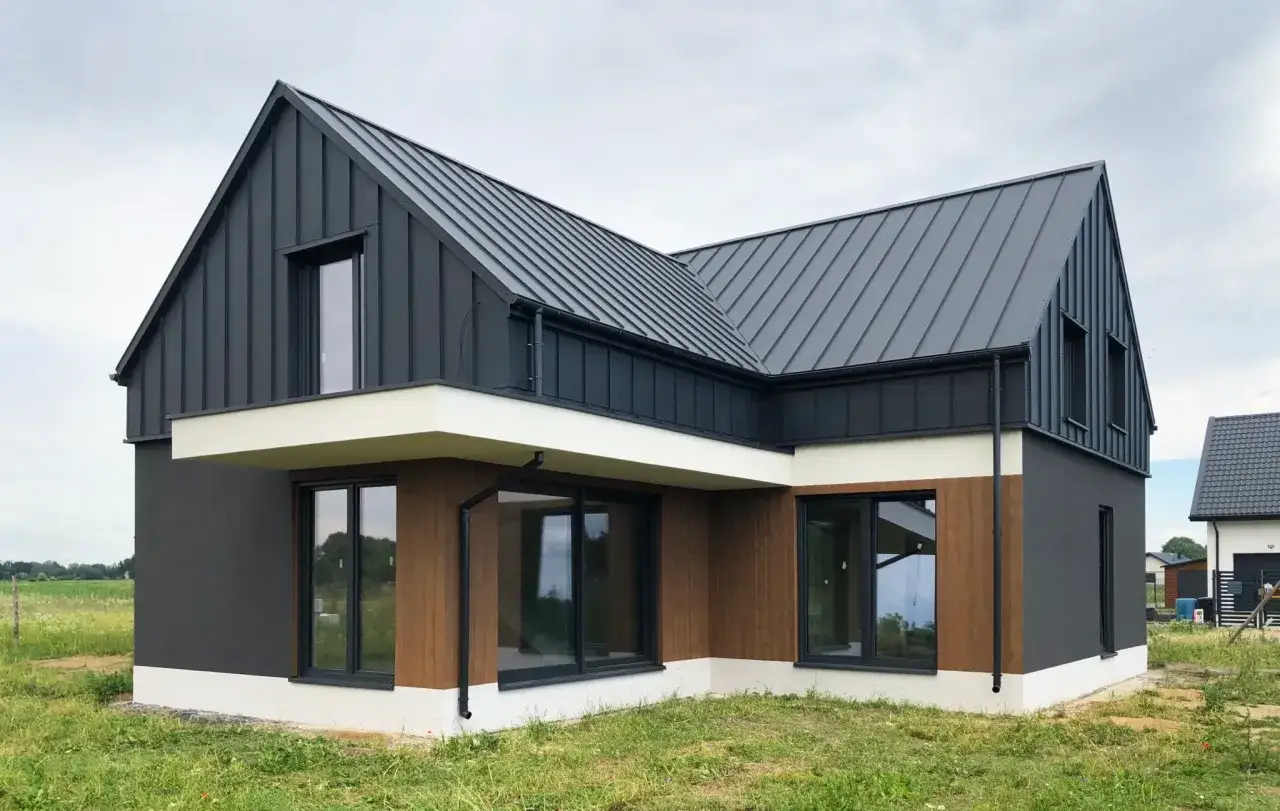 Nowoczesny dom w stanie deweloperskim, z czarnym, metalowym dachem i grafitową elewacją, uzupełnioną drewnianymi panelami.
