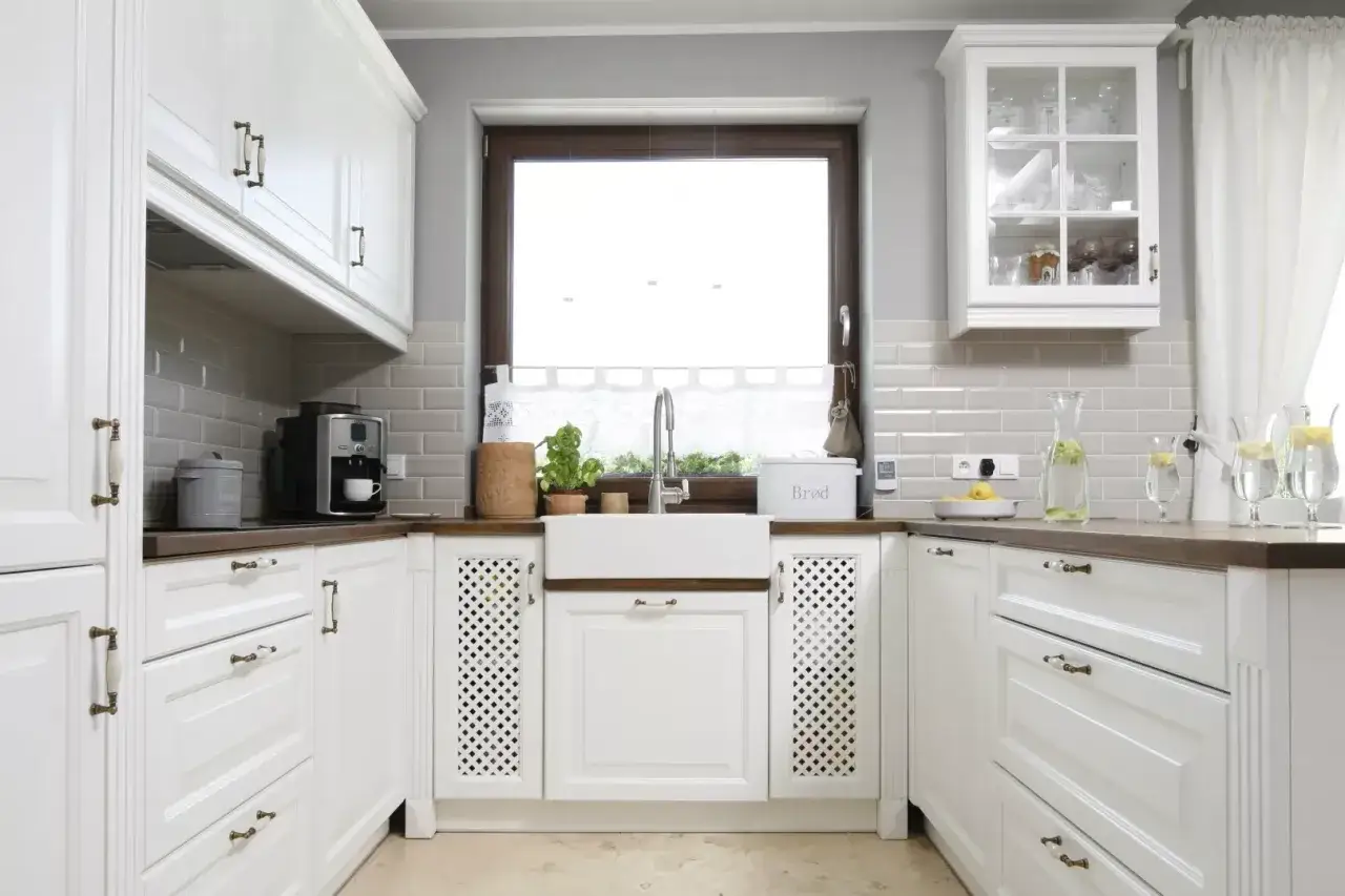 Jasna kuchnia z białymi meblami i drewnianym blatem. Okno w kuchni znajduje się nad białym zlewozmywakiem, na wygodnej wysokości.