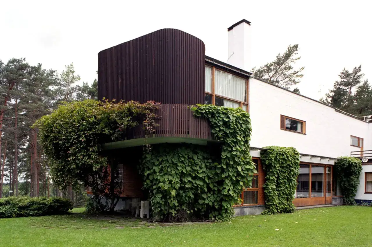 Villa Mairea, modernistyczna willa otoczona lasem, z charakterystyczną, zaokrągloną fasadą i bujną zielenią pnącą się po ścianach.