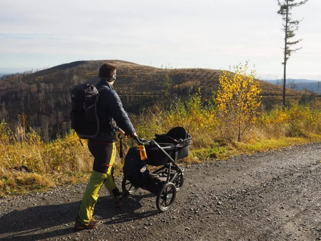Wędr&oacute;wka gdzie w g&oacute;ry z dzieckiem w w&oacute;zku. Jesienny krajobraz, osoba z plecakiem pcha w&oacute;zek po kamienistej drodze.