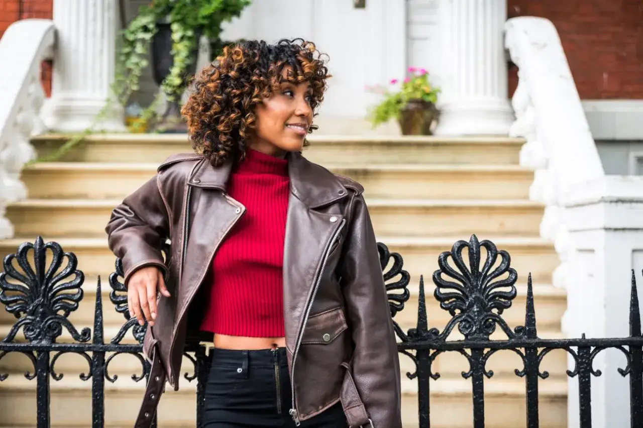 Kobieta w brązowej, błyszczącej kurtce motocyklowej i czerwonym golfie. Czy błyszczące kurtki są modne? Ta stylizacja sugeruje, że tak!