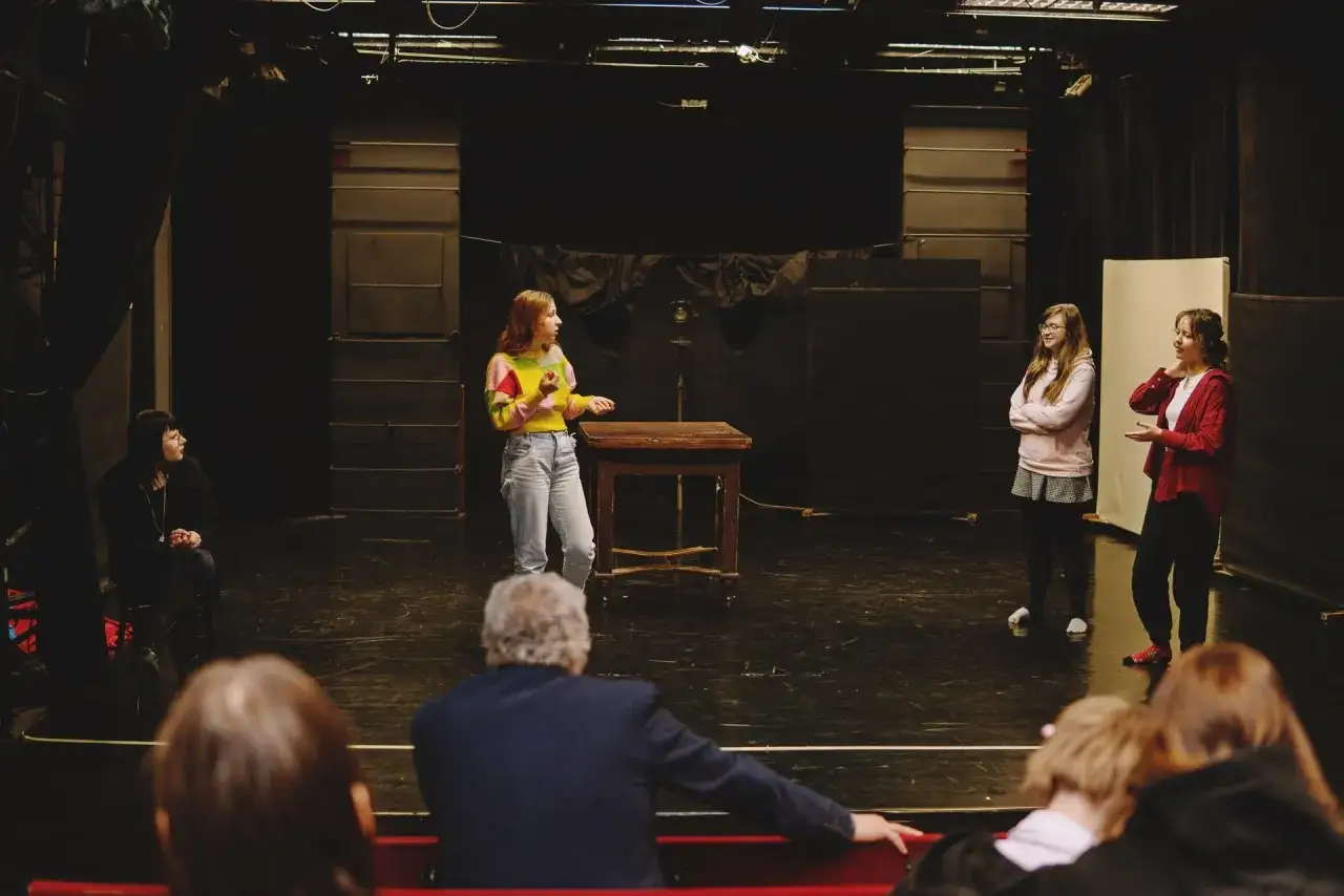 Młodzi aktorzy ćwiczą scenę w teatrze dla nastolatków. Na scenie stoi drewniany stolik, a w tle widać kurtyny i reflektory.