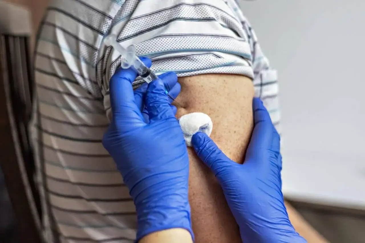 Ręka w niebieskiej rękawiczce podaje zastrzyk domięśniowy w ramię. Po zastrzyku może pojawić się niewielkie zgrubienie.