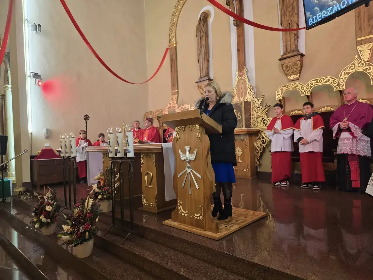 Kobieta przemawia przy ambionie w kościele. W tle widać ministrantów i biskupa. Zastanawiasz się, ile kosztuje bierzmowanie dla dorosłych?