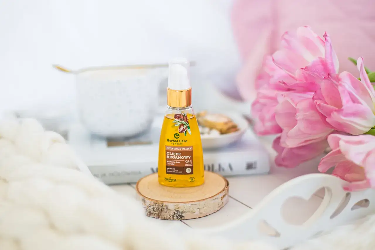 Odkryj, jak używać olejku arganowego do włosów, skóry i paznokci. Flakonik na drewnianym krążku, obok różowych tulipanów.