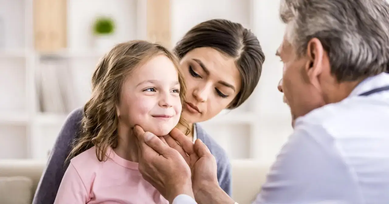 Lekarz bada gardło dziewczynki, zaniepokojona mama czuwa obok. To może być zapalenie oskrzeli u dzieci.