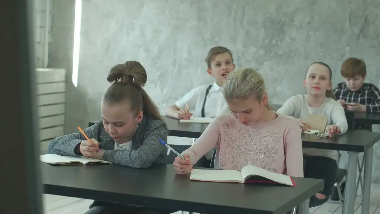 Dzieci piszą w zeszytach, podczas gdy chłopiec z tyłu rozmawia. Sytuacja, gdy szkoła nie realizuje zaleceń poradni, może prowadzić do takich rozproszeń.