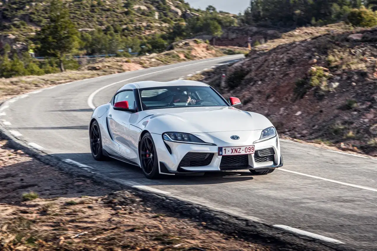 Biała Toyota Supra z czerwonymi lusterkami pędzi po krętej drodze. Zastanawiasz się, ile kosztuje Toyota Supra?