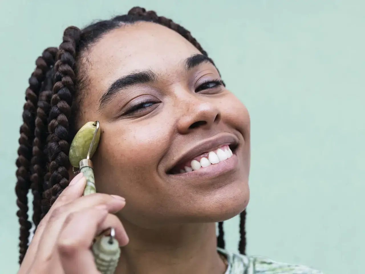 Uśmiechnięta kobieta z warkoczami używa jadeitowego roller do twarzy jak używać, masując policzek.