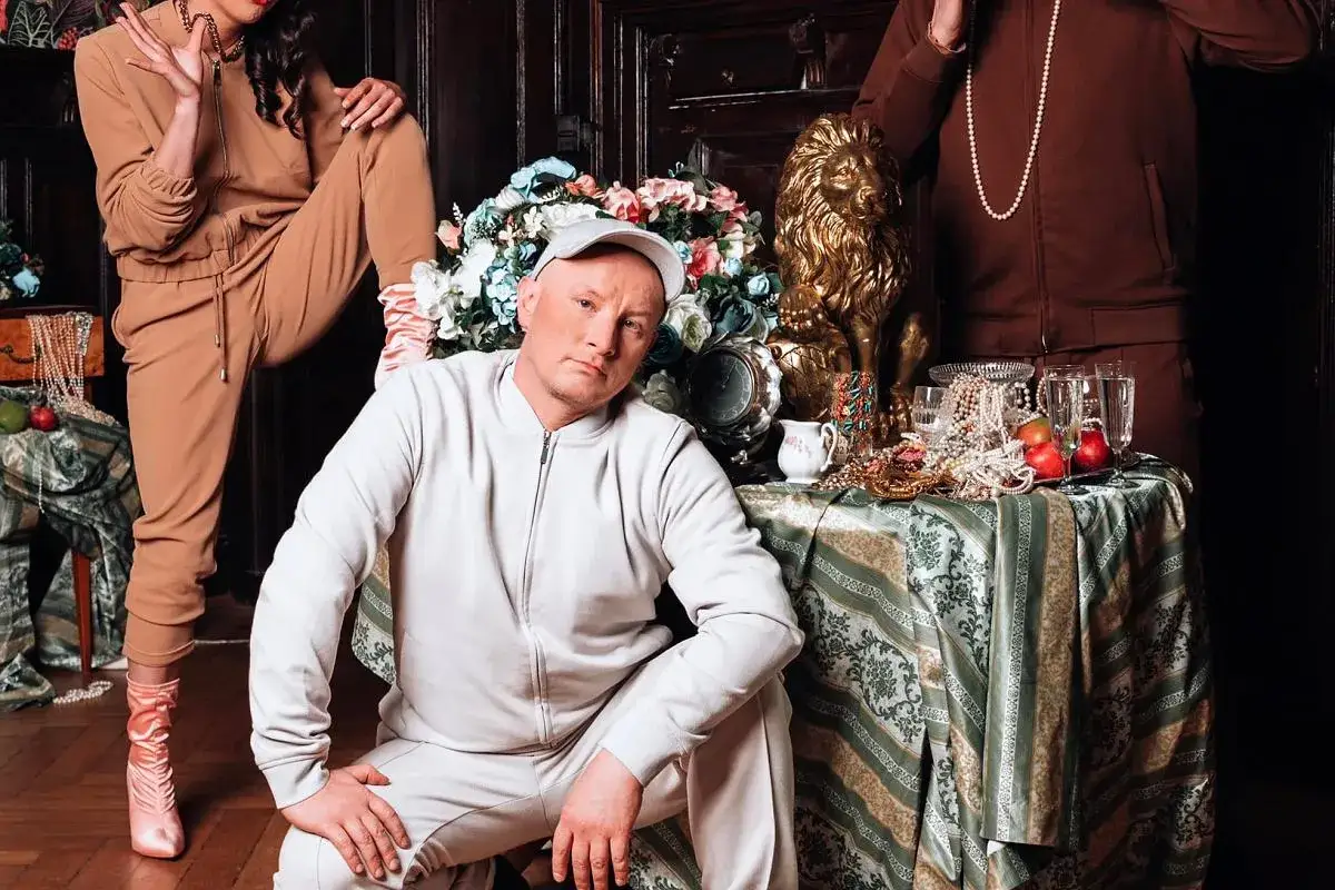 Kabaret K2 skład: mężczyzna w jasnym dresie, kobieta w beżowym dresie i druga osoba w brązowym dresie, otoczeni bogactwem i kwiatami.