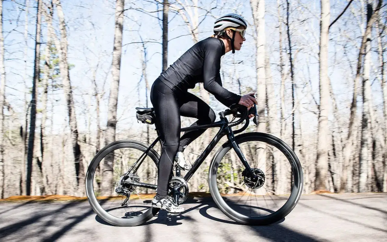 Kobieta w czarnym stroju kolarskim i kasku jedzie rowerem po leśnej drodze. Idealny przykład, jak się ubrać na rower w chłodniejszy dzień.