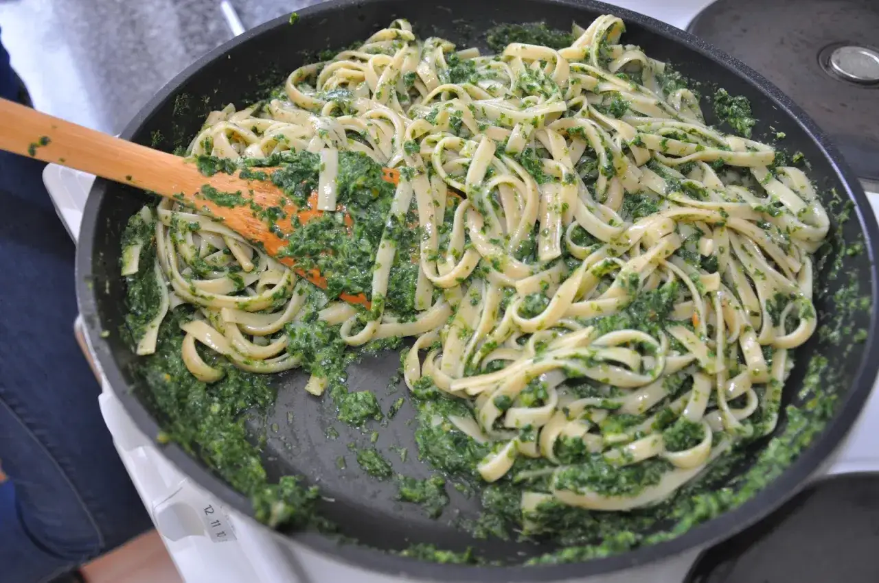 Makaron tagliatelle z zielonym pesto ze słoika, mieszany drewnianą łopatką na patelni.
