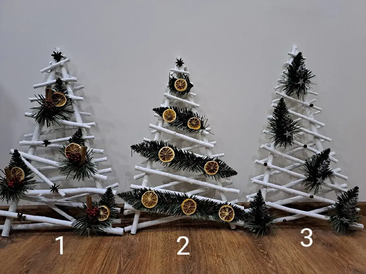 Jak zrobić choinkę z drewna? Trzy ozdobne, białe choinki z gałązek, udekorowane suszonymi pomarańczami, szyszkami i cynamonem.